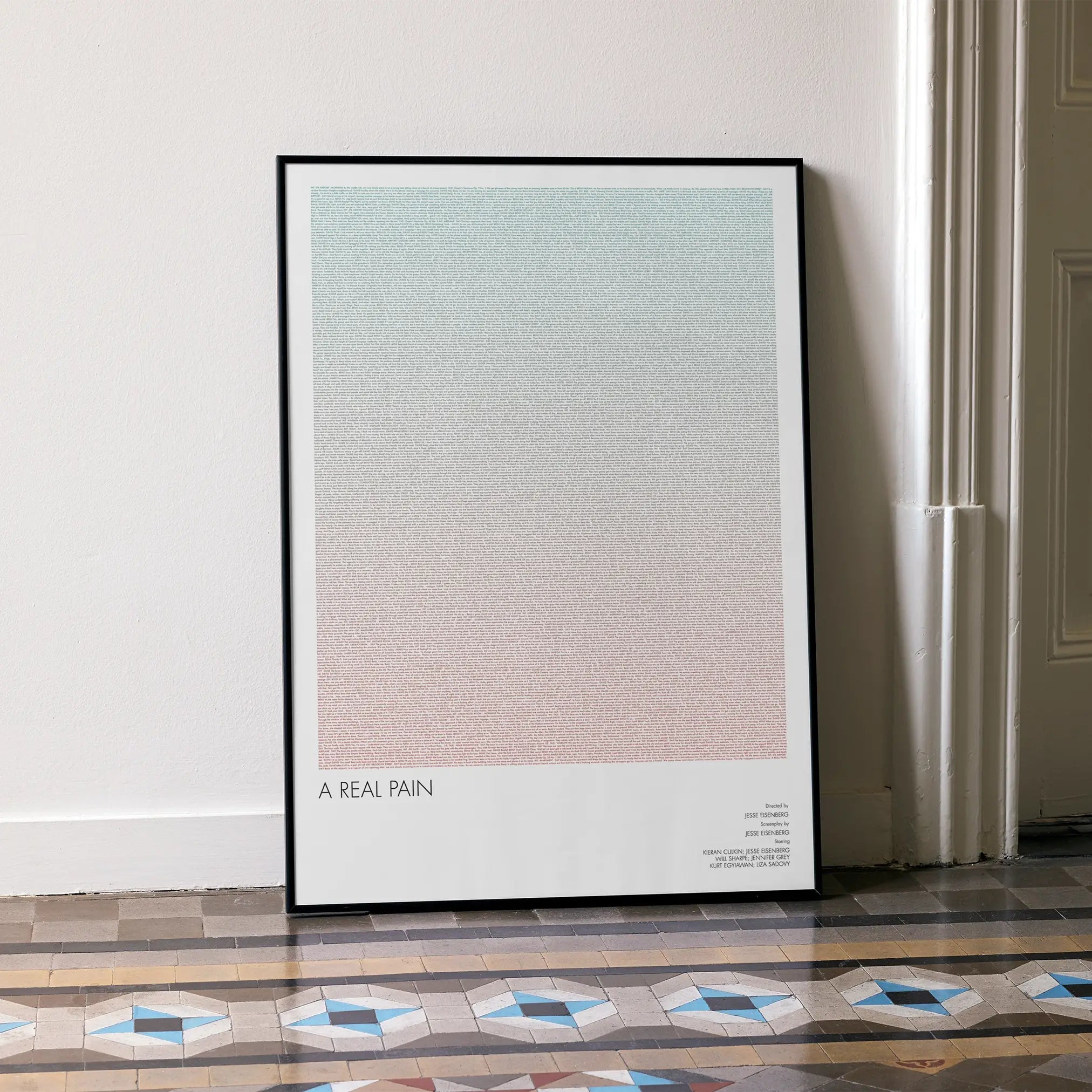 A framed A Real Pain movie script poster leaning against a wall in a minimalist interior.