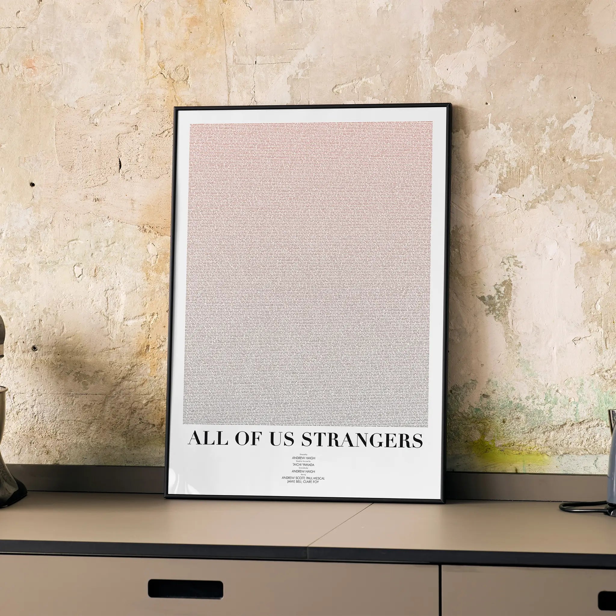 A framed All of Us Strangers movie script poster displayed on a cabinet in a minimalist interior.