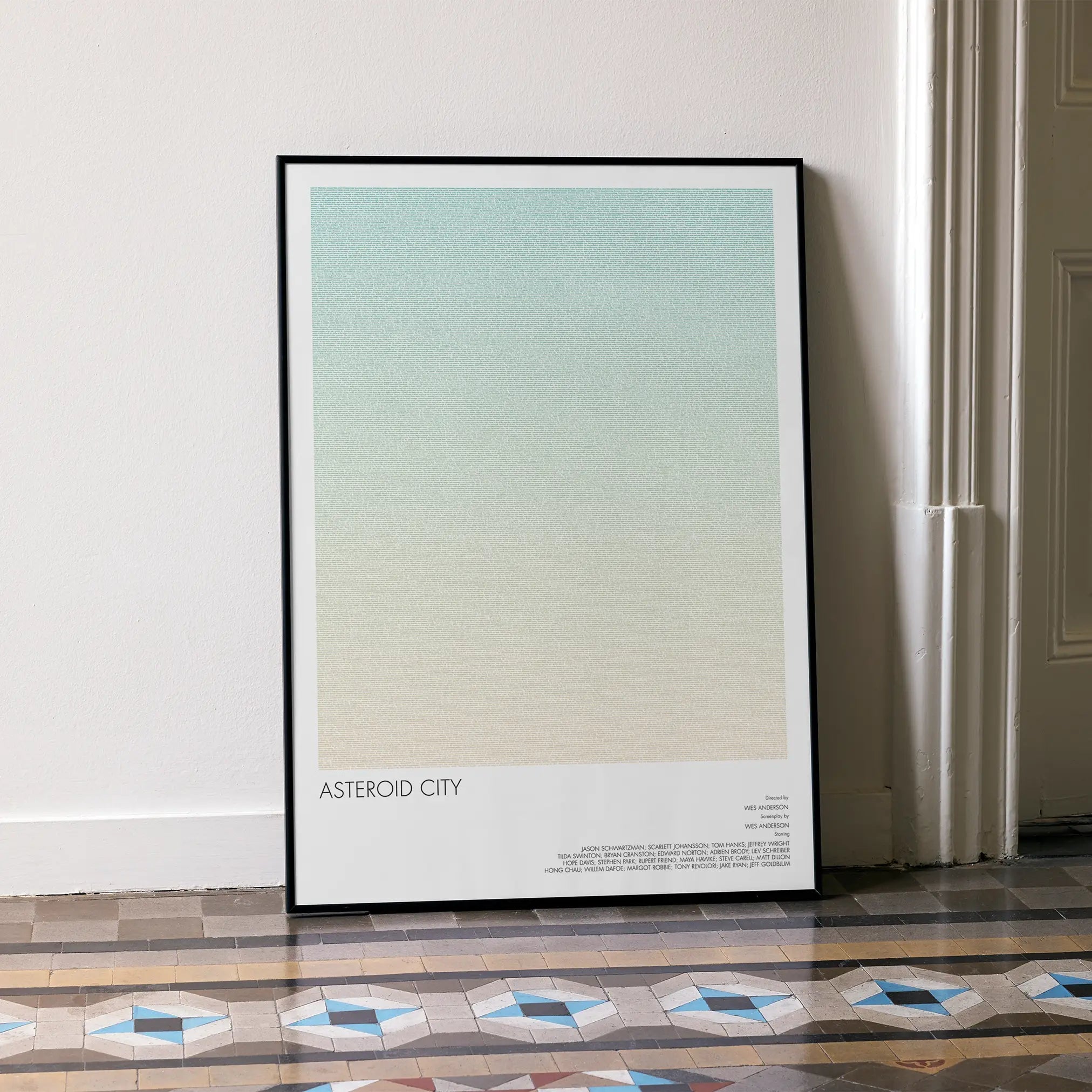 A framed Asteroid City movie script poster leaning against a wall in a minimalist interior.