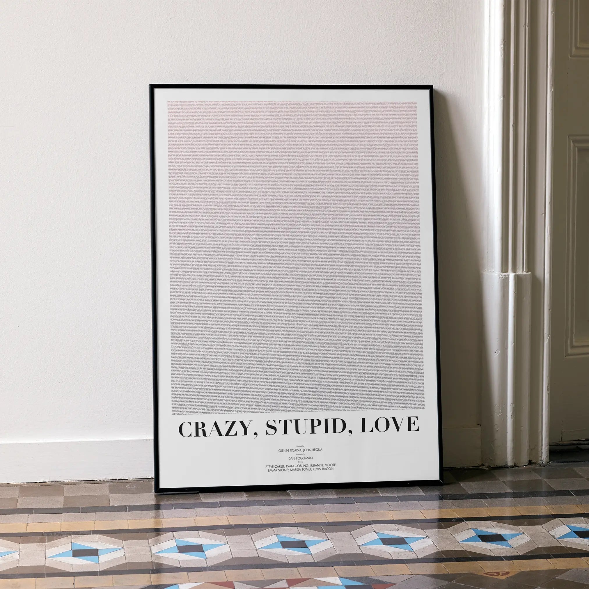 A framed Crazy, Stupid, Love. movie script poster leaning against a wall in a minimalist interior.