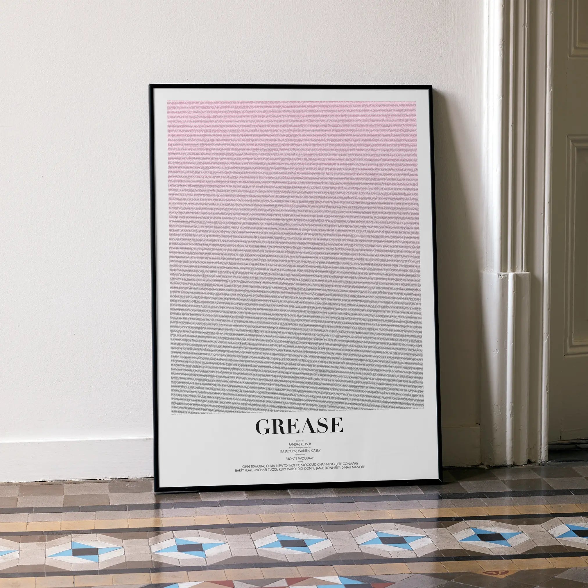 A framed Grease movie script poster leaning against a wall in a minimalist interior.