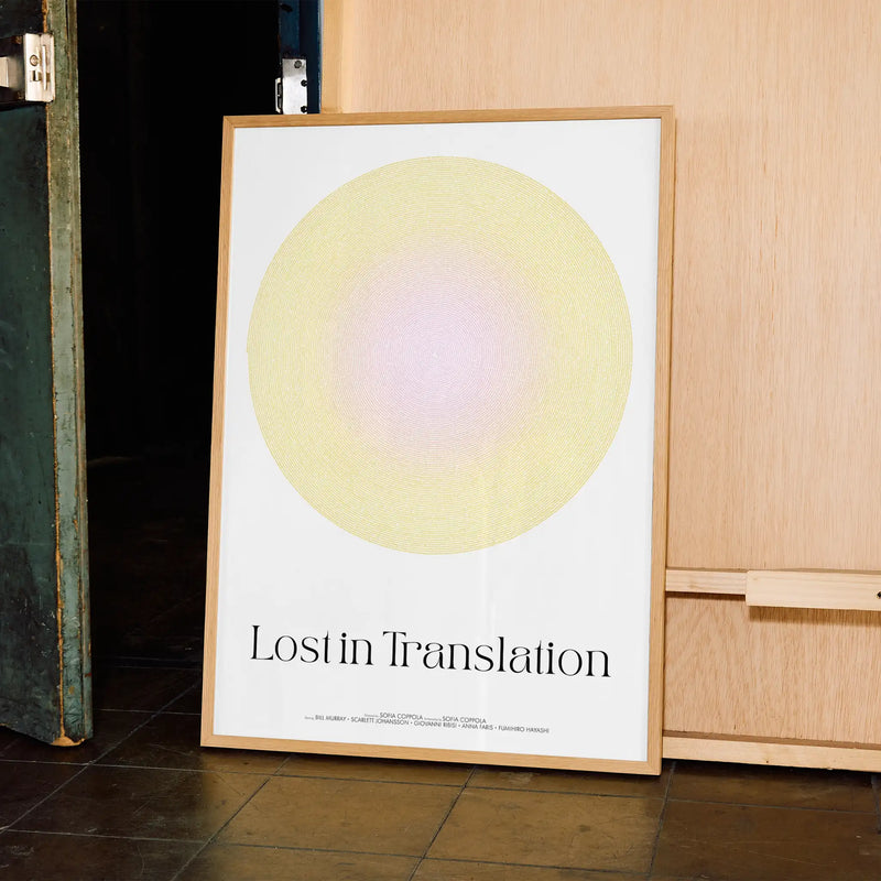 Framed Lost in Translation movie poster with a yellow and pink screenplay text circle and 'Lost in Translation' text on a white background, leaning against a wooden wall.