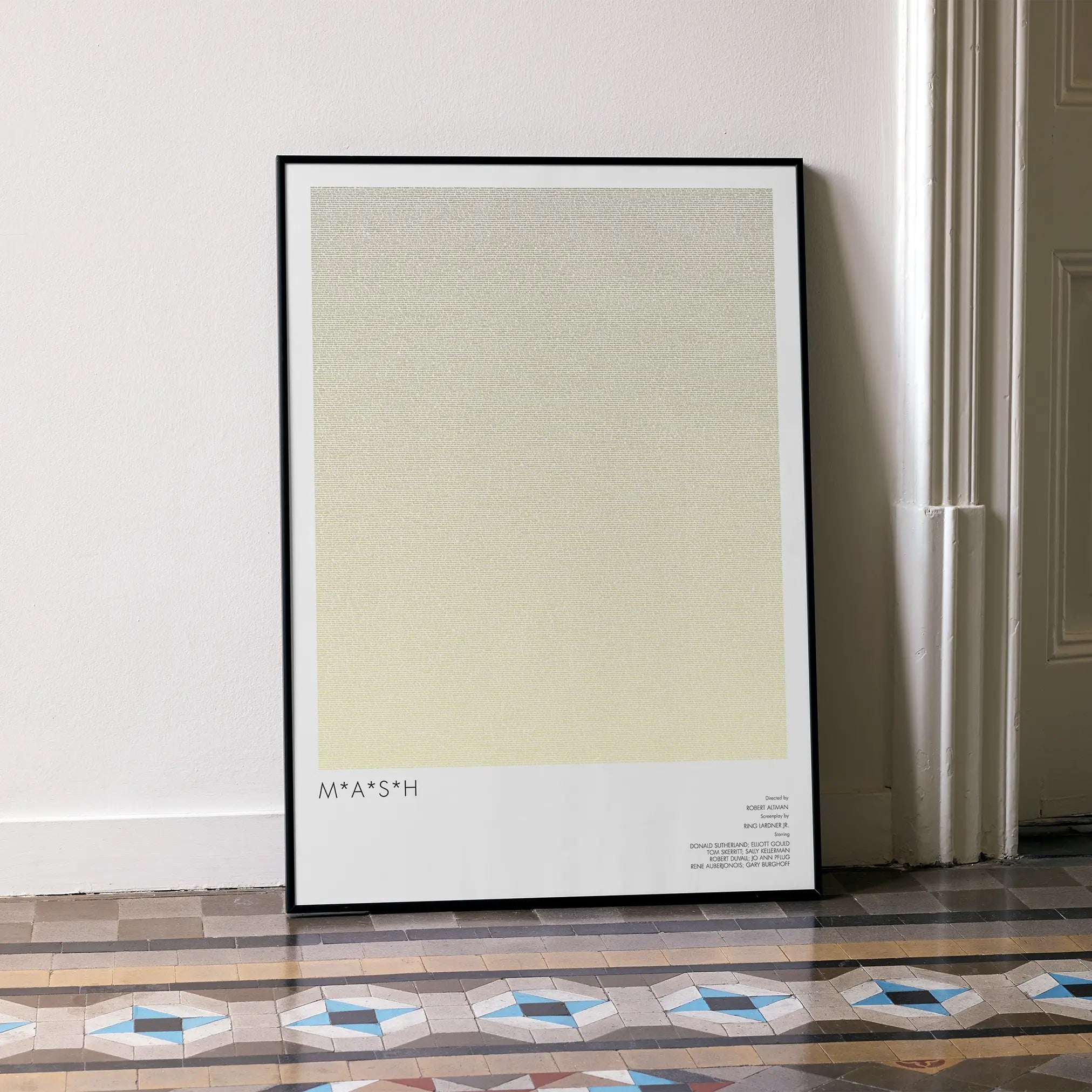 A framed M*A*S*H movie script poster leaning against a wall in a minimalist interior.