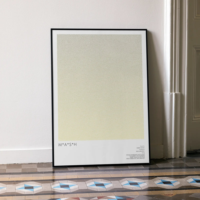 A framed M*A*S*H movie script poster leaning against a wall in a minimalist interior.