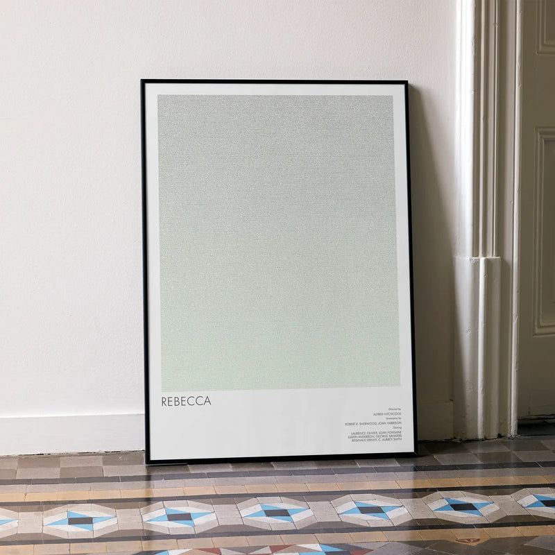 A framed movie poster for Hitchcock's "Rebecca", leans against a white wall.