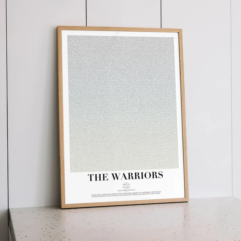A framed minimalist poster featuring a large light gray textured field with the words 'THE WARRIORS' in black serif text at the bottom, displayed against a white paneled wall on a light speckled surface.